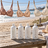 Four white bottles of Forever Now body wash arranged on a weathered wooden table at the beach, with lace bras hanging on a clothesline in the background and the ocean and shoreline visible.