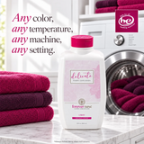 A bottle of Forever New Delicate Fabric Care Wash in orange-scented liquid is displayed on a marble countertop in a bright laundry room. The white bottle has a pink cap and a pink floral label.